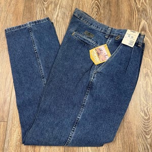 May include: Blue denim Wrangler jeans with a straight leg cut. The jeans have a classic five-pocket design, a zipper fly, and a Wrangler tag. The jeans are folded on a wooden surface.