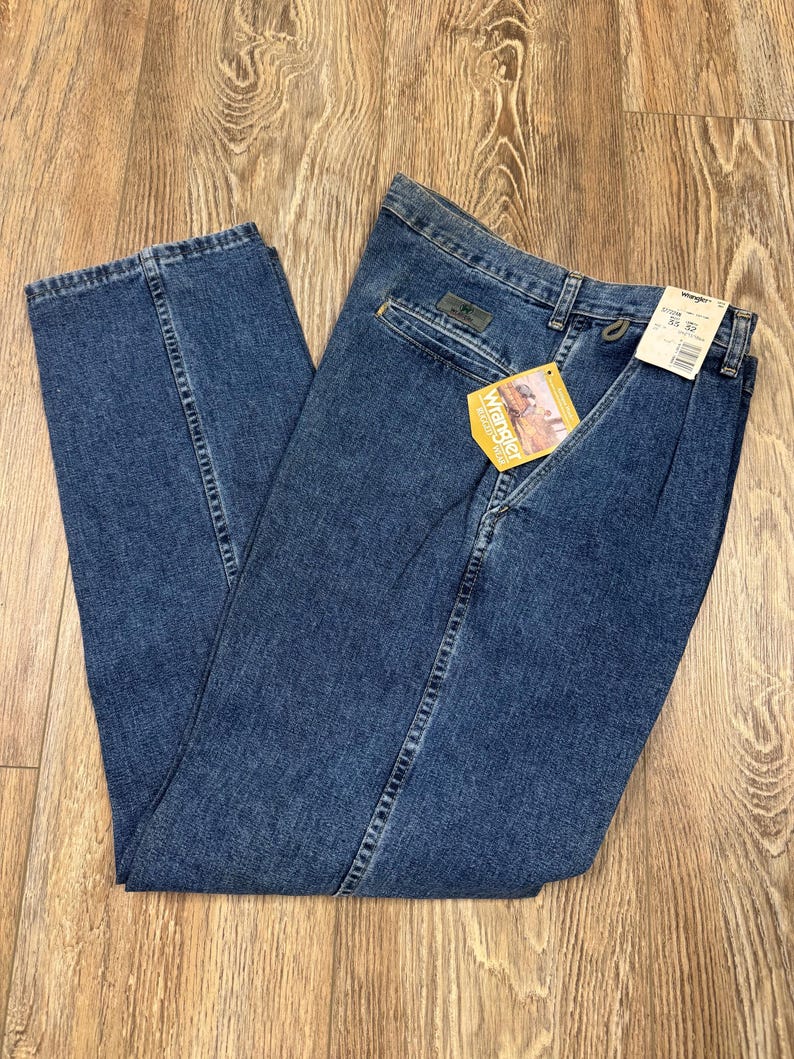May include: Blue denim Wrangler jeans with a straight leg cut. The jeans have a classic five-pocket design, a zipper fly, and a Wrangler tag. The jeans are folded on a wooden surface.