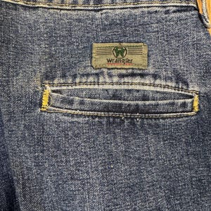 May include: Close-up of the back pocket of blue denim jeans. The pocket has yellow stitching and a rectangular label with the brand name "Wrangler" in white text. The label is green and tan.