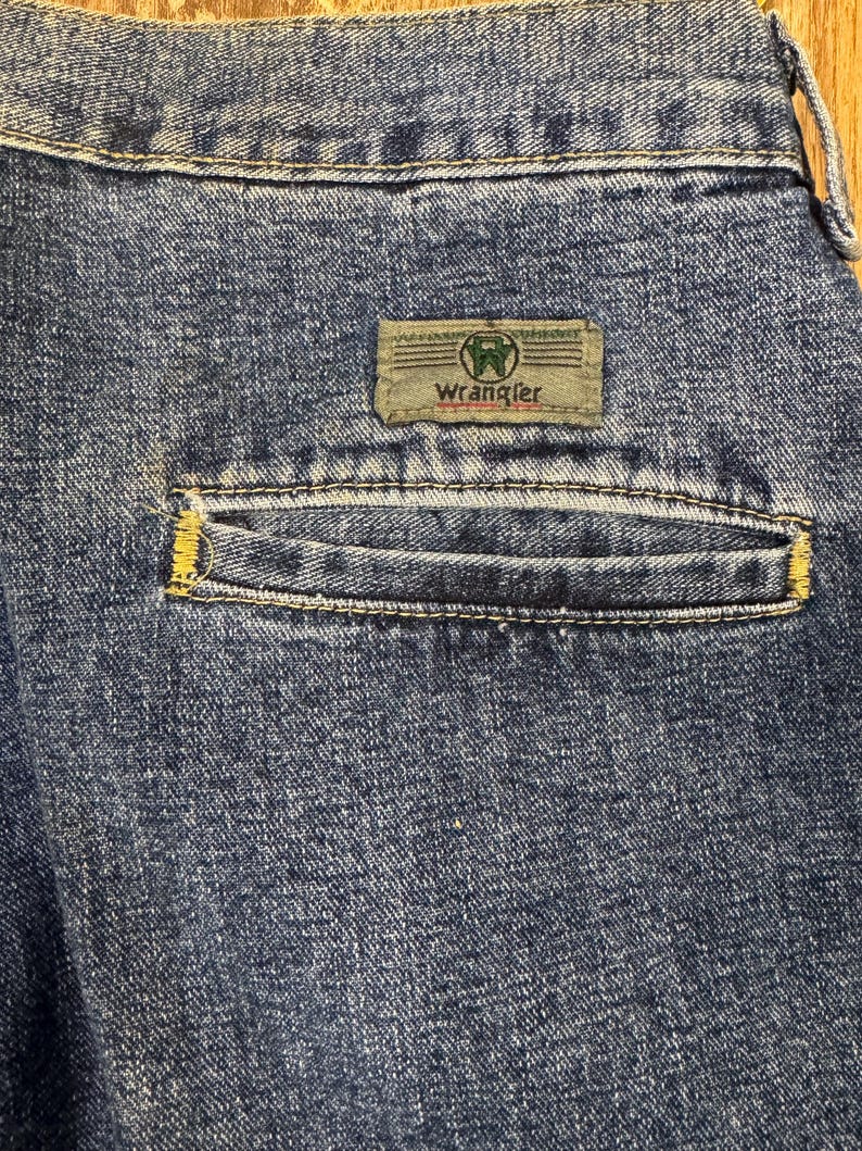 May include: Close-up of the back pocket of blue denim jeans. The pocket has yellow stitching and a rectangular label with the brand name "Wrangler" in white text. The label is green and tan.