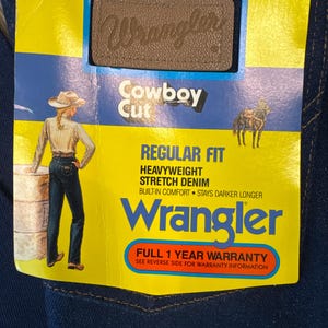 May include: A close-up of a pair of dark blue Wrangler Cowboy Cut jeans. The jeans have a yellow tag with the Wrangler logo, the words "STRETCH DENIM", and "REGULAR FIT". The tag also features an illustration of a person in a cowboy hat.