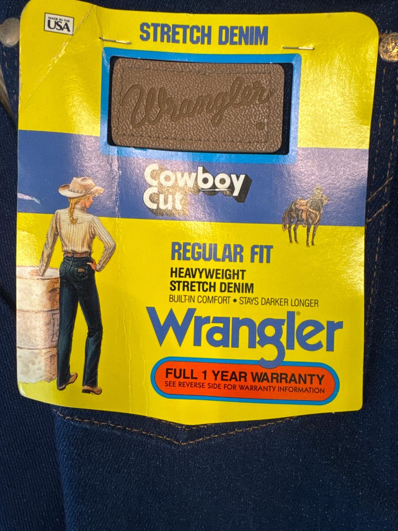May include: A close-up of a pair of dark blue Wrangler Cowboy Cut jeans. The jeans have a yellow tag with the Wrangler logo, the words "STRETCH DENIM", and "REGULAR FIT". The tag also features an illustration of a person in a cowboy hat.