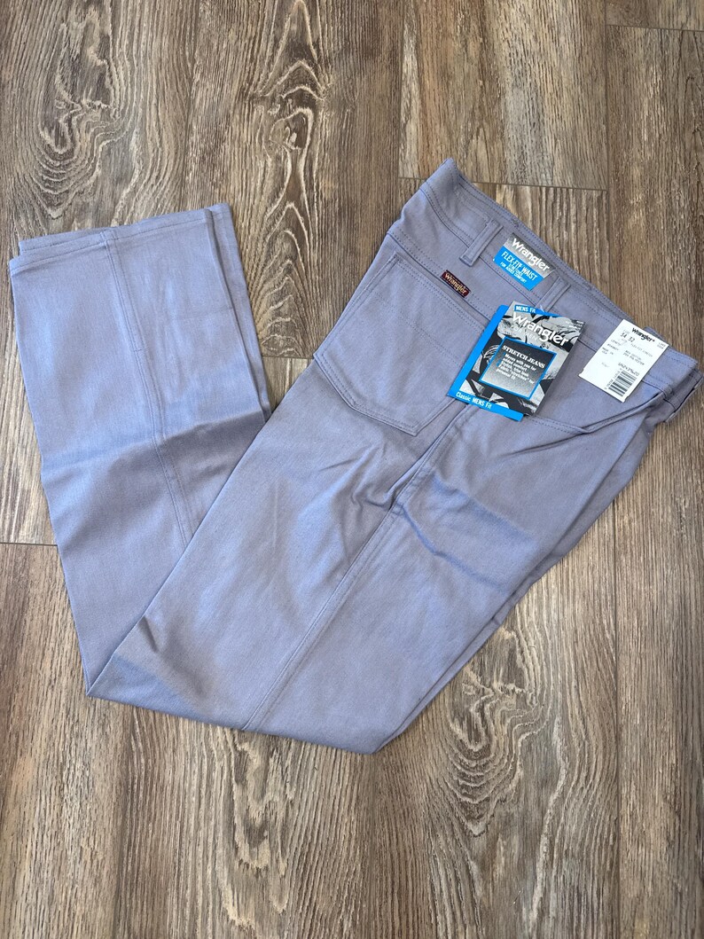 May include: Light blue Wrangler Flex Fit waist stretch jeans. The jeans have a classic fit and feature a tag with the Wrangler logo and product information. The pants are laid out on a wooden surface.