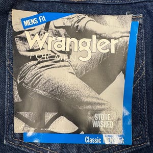 K&ouml;nnte beinhalten: Nahaufnahme einer blauen Jeans mit einem Wrangler-Etikett auf der Ges&auml;&szlig;tasche. Das Etikett lautet "Wrangler FOR MEN" und "STONE WASHED". Die Jeans hat eine klassische Herrenpassform.