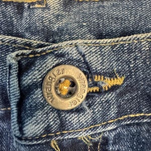 May include: Close-up of a blue denim jeans button with the brand name "Wrangler" embossed on it. The button is silver-coloured and has a yellow centre. The jeans fabric is a dark blue wash with visible stitching.
