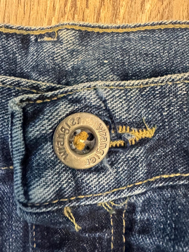 May include: Close-up of a blue denim jeans button with the brand name "Wrangler" embossed on it. The button is silver-coloured and has a yellow centre. The jeans fabric is a dark blue wash with visible stitching.