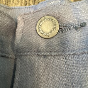 May include: Close-up of a light blue denim button with the word "Wrangler" around the edge. The button is attached to a light blue denim garment. The button is silver in color.
