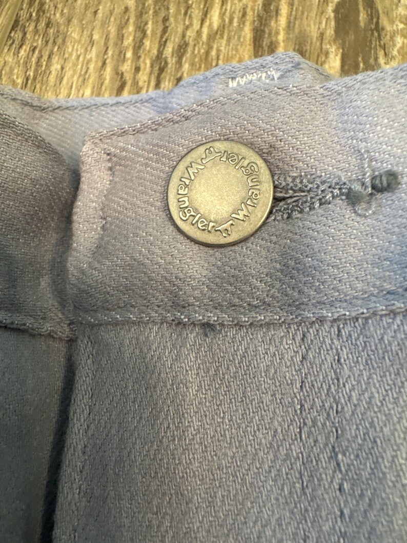 May include: Close-up of a light blue denim button with the word "Wrangler" around the edge. The button is attached to a light blue denim garment. The button is silver in color.