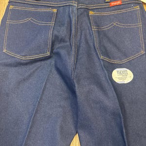 May include: Close-up of the back of dark blue denim jeans with yellow stitching. The jeans have two back pockets with a unique curved design. The Wrangler logo is visible on the back pocket. A Flex Fit tag is also visible.