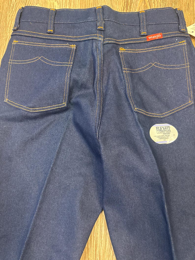 May include: Close-up of the back of dark blue denim jeans with yellow stitching. The jeans have two back pockets with a unique curved design. The Wrangler logo is visible on the back pocket. A Flex Fit tag is also visible.