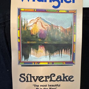 K&ouml;nnte beinhalten: Ein braunes Etikett mit dem Text "Wrangler" in blauen Buchstaben. Das Etikett zeigt eine farbenfrohe Illustration einer Bergsee-Szene mit einem Berg im Hintergrund. Der Text "SilverLake" ist in schwarzen Buchstaben unter dem Bild gedruckt. Der Text "The most beautiful fit in the West" ist in schwarzen Buchstaben unter dem Text "SilverLake" gedruckt.