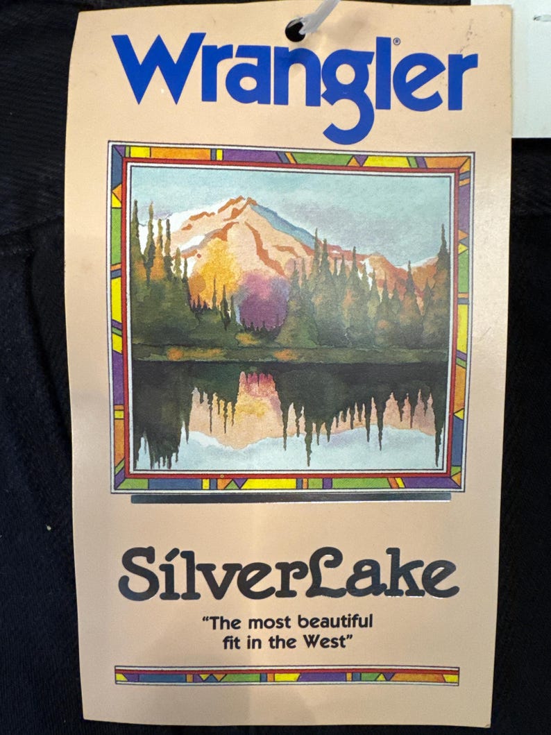 K&ouml;nnte beinhalten: Ein braunes Etikett mit dem Text "Wrangler" in blauen Buchstaben. Das Etikett zeigt eine farbenfrohe Illustration einer Bergsee-Szene mit einem Berg im Hintergrund. Der Text "SilverLake" ist in schwarzen Buchstaben unter dem Bild gedruckt. Der Text "The most beautiful fit in the West" ist in schwarzen Buchstaben unter dem Text "SilverLake" gedruckt.