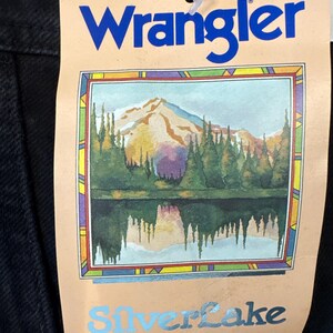 May include: A black garment tag with the blue word "Wrangler" at the top. Below is a colourful illustration of a mountain range, trees, and a lake. The word "Silverlake" is below the illustration, with the text "The most beautiful fit in the West."