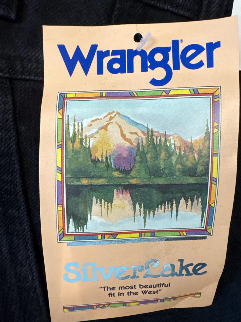 May include: A black garment tag with the blue word "Wrangler" at the top. Below is a colourful illustration of a mountain range, trees, and a lake. The word "Silverlake" is below the illustration, with the text "The most beautiful fit in the West."