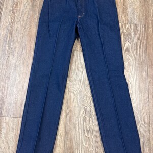 May include: Dark blue denim jeans with a classic five-pocket design. The jeans have a straight leg cut and a button and zipper closure. The fabric appears to be a medium-weight denim. The jeans are new with a tag attached.