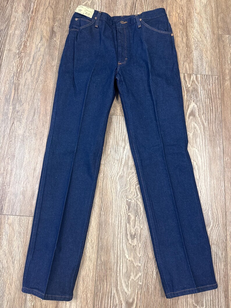 May include: Dark blue denim jeans with a classic five-pocket design. The jeans have a straight leg cut and a button and zipper closure. The fabric appears to be a medium-weight denim. The jeans are new with a tag attached.