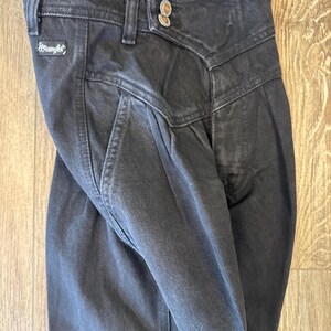 May include: Close-up of black denim Wrangler jeans. The jeans feature a high waist, pleats, and a button closure. The Wrangler logo is visible on the front pocket. The jeans are folded, showing the texture of the denim.