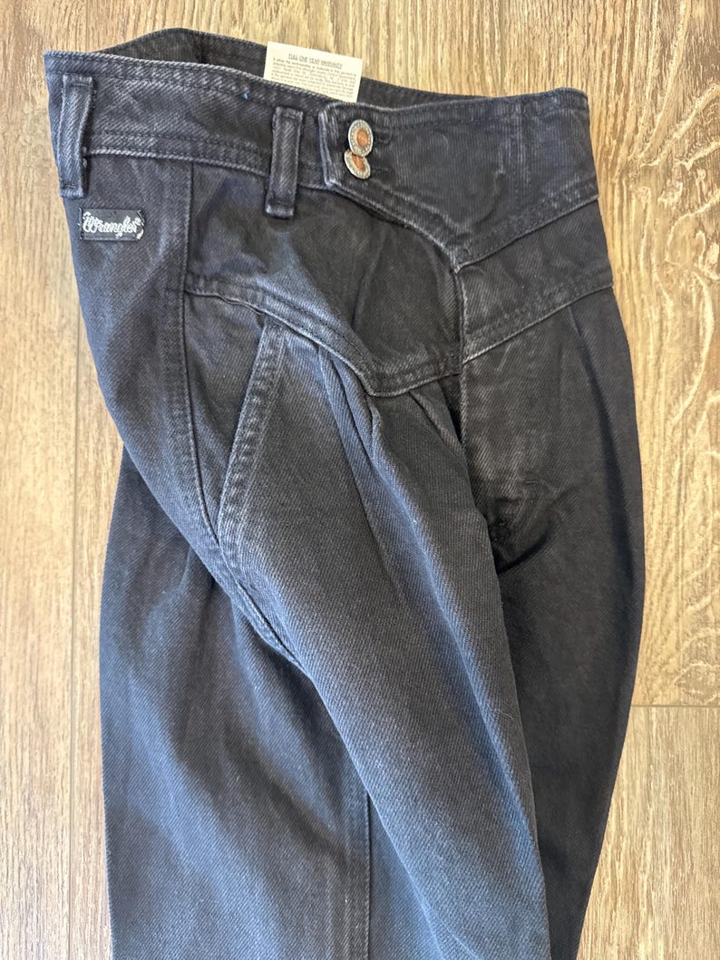 May include: Close-up of black denim Wrangler jeans. The jeans feature a high waist, pleats, and a button closure. The Wrangler logo is visible on the front pocket. The jeans are folded, showing the texture of the denim.