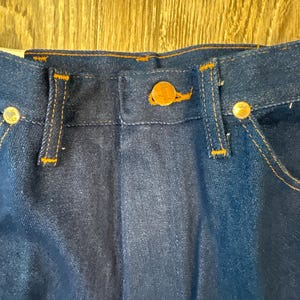 May include: Close-up of the waistband and button closure of dark blue denim jeans. The jeans have orange stitching and gold-colored metal buttons. The button closure features a gold-colored button with a logo.