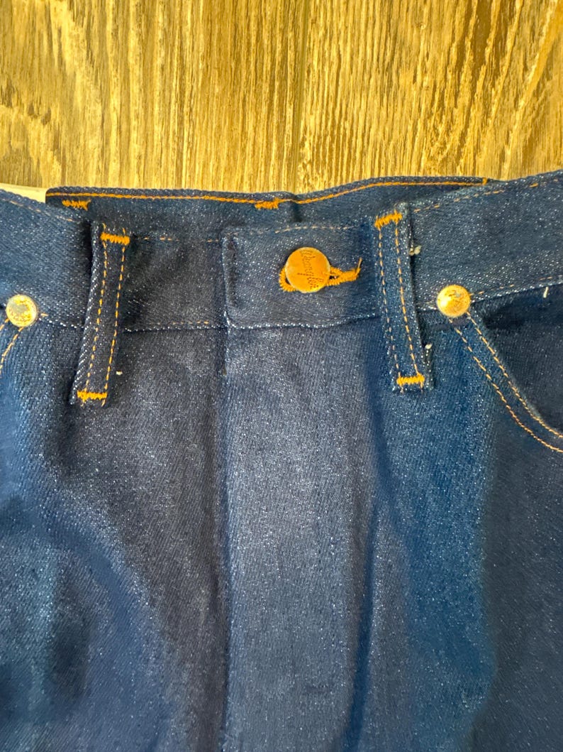 May include: Close-up of the waistband and button closure of dark blue denim jeans. The jeans have orange stitching and gold-colored metal buttons. The button closure features a gold-colored button with a logo.