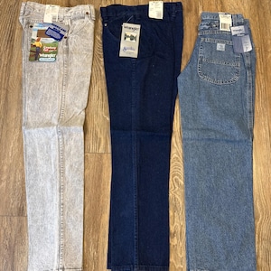 May include: Three pairs of Wrangler jeans in different washes: a light gray acid wash, a dark blue, and a medium blue. Each pair has tags attached. The jeans are laid out on a wooden surface.