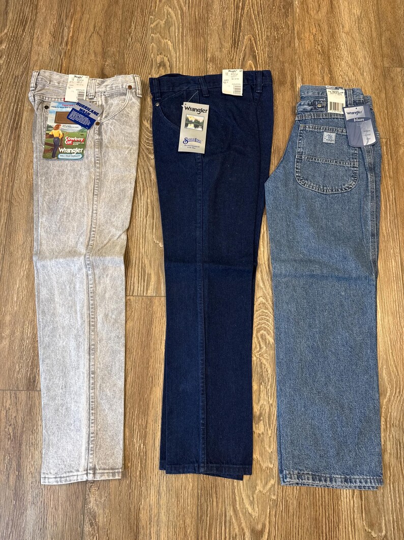 May include: Three pairs of Wrangler jeans in different washes: a light gray acid wash, a dark blue, and a medium blue. Each pair has tags attached. The jeans are laid out on a wooden surface.