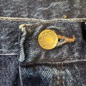 May include: Close-up of dark blue denim jeans. A brass button with the word "Wrangler" embossed around the edge is visible. The jean's stitching is present, and the fabric shows signs of wear.