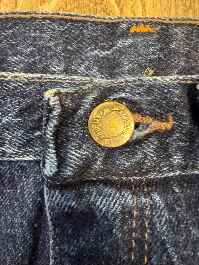 May include: Close-up of dark blue denim jeans. A brass button with the word "Wrangler" embossed around the edge is visible. The jean's stitching is present, and the fabric shows signs of wear.
