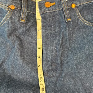 May include: A close-up of a pair of blue denim jeans with a brown leather label that reads "Wrangler" and a measuring tape showing the waist measurement is 32 inches.