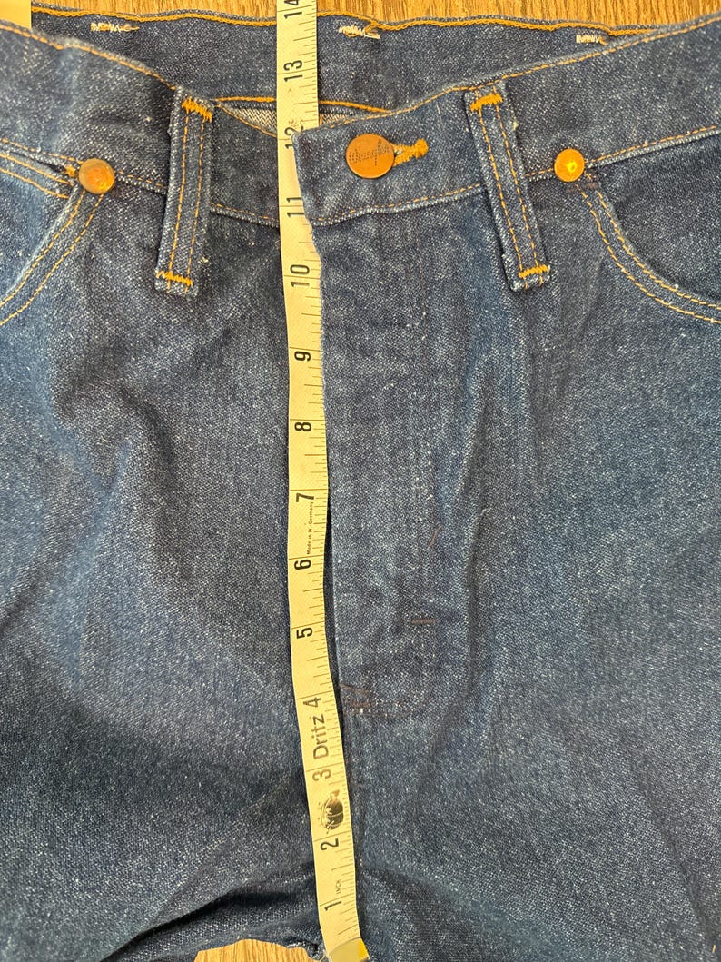 May include: A close-up of a pair of blue denim jeans with a brown leather label that reads "Wrangler" and a measuring tape showing the waist measurement is 32 inches.