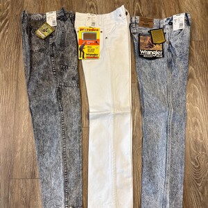 K&ouml;nnte beinhalten: Drei Jeanshosen von Wrangler. Eine Hose ist wei&szlig;, die anderen beiden sind s&auml;uregewaschen. Jede Hose hat ein Etikett. Die Jeans werden auf einer Holzoberfl&auml;che pr&auml;sentiert.