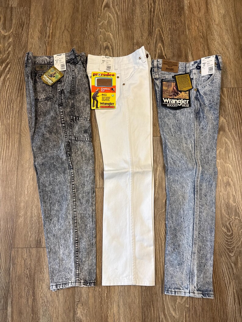 K&ouml;nnte beinhalten: Drei Jeanshosen von Wrangler. Eine Hose ist wei&szlig;, die anderen beiden sind s&auml;uregewaschen. Jede Hose hat ein Etikett. Die Jeans werden auf einer Holzoberfl&auml;che pr&auml;sentiert.