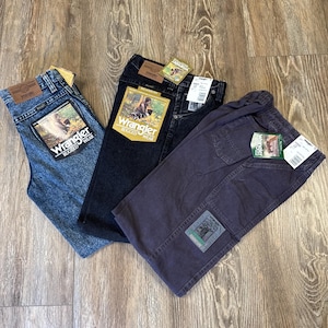 Może przedstawiać: Trzy pary dżins&oacute;w Wrangler w r&oacute;żnych kolorach: jasnoniebieskim, czarnym i ciemnofioletowym. Każda para ma metkę z logo Wrangler i grafiką osoby na zewnątrz. Dżinsy są złożone i leżą na drewnianej powierzchni.
