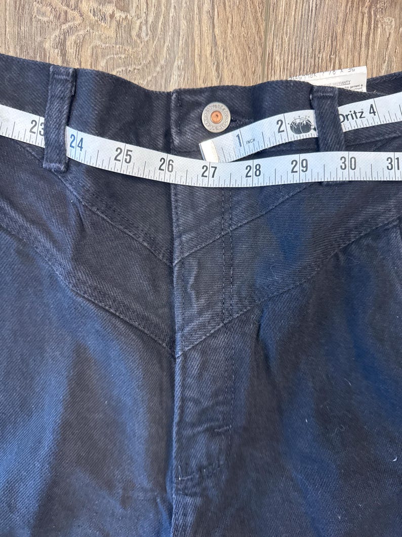 K&ouml;nnte beinhalten: Schwarze Jeans aus Denim mit hoher Taille und geradem Bein. Die Jeans hat ein einzigartiges Design mit N&auml;hten an den vorderen Taschen und einem Knopfverschluss. Die Taille wird mit einem Ma&szlig;band gemessen und zeigt eine Gr&ouml;&szlig;e von 68,58 cm.