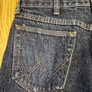 May include: Detail of dark blue denim jeans, focusing on the back pocket with tan 'W' stitching. The jeans show classic design elements, including a reinforced waistband and visible stitching.