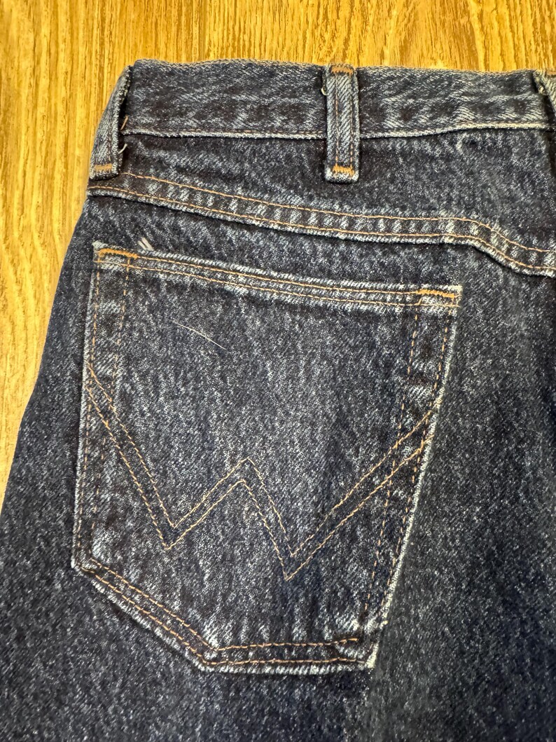 May include: Detail of dark blue denim jeans, focusing on the back pocket with tan 'W' stitching. The jeans show classic design elements, including a reinforced waistband and visible stitching.