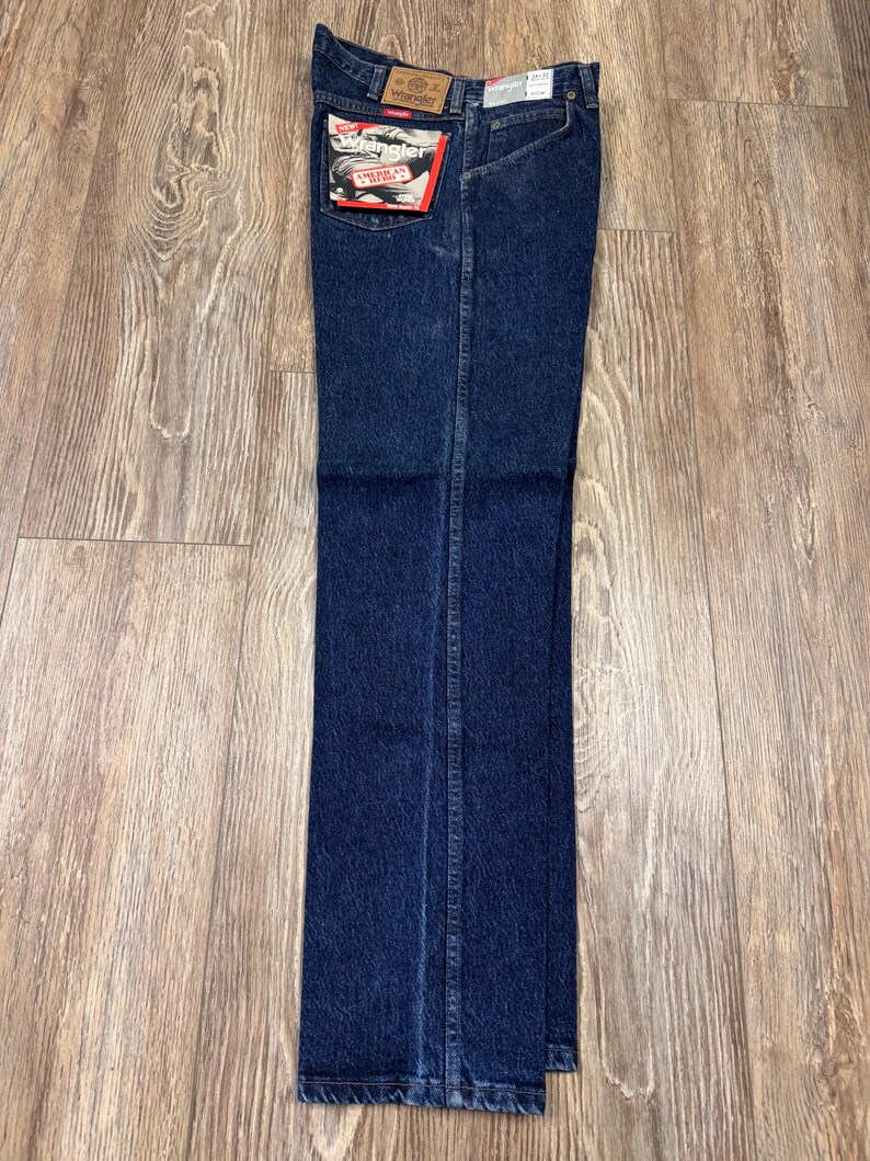 May include: Dark blue denim jeans with a classic straight leg fit. The jeans have a Wrangler brand label on the back pocket and a brown leather patch at the waistband. The jeans are new with tags.
