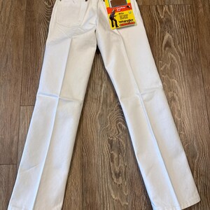 K&ouml;nnte beinhalten: Wei&szlig;e Wrangler ProRodeo Jeans mit geradem Bein. Die Jeans haben ein klassisches F&uuml;nf-Taschen-Design und ein Wrangler-Etikett. Auf dem Etikett steht "Cowboy Cut" und "Boys". Die Jeans liegen flach auf einer Holzoberfl&auml;che.