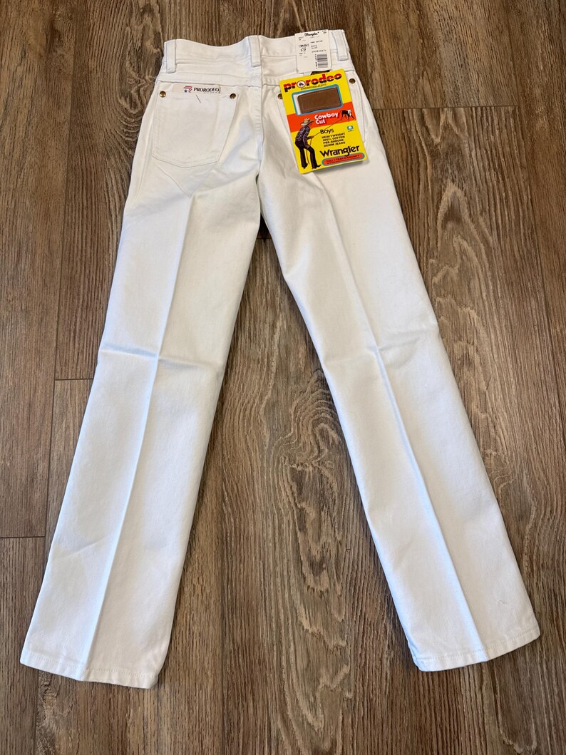 K&ouml;nnte beinhalten: Wei&szlig;e Wrangler ProRodeo Jeans mit geradem Bein. Die Jeans haben ein klassisches F&uuml;nf-Taschen-Design und ein Wrangler-Etikett. Auf dem Etikett steht "Cowboy Cut" und "Boys". Die Jeans liegen flach auf einer Holzoberfl&auml;che.