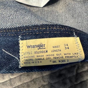 K&ouml;nnte beinhalten: Nahaufnahme eines Jeansetiketts der Marke Wrangler. Das gelbe Etikett mit schwarzem Text zeigt eine Bundweite von 86 cm und eine L&auml;nge von 86 cm an. Das Etikett enth&auml;lt auch Waschanweisungen und die Modellnummer 85200SW. Die Jeans ist blau.