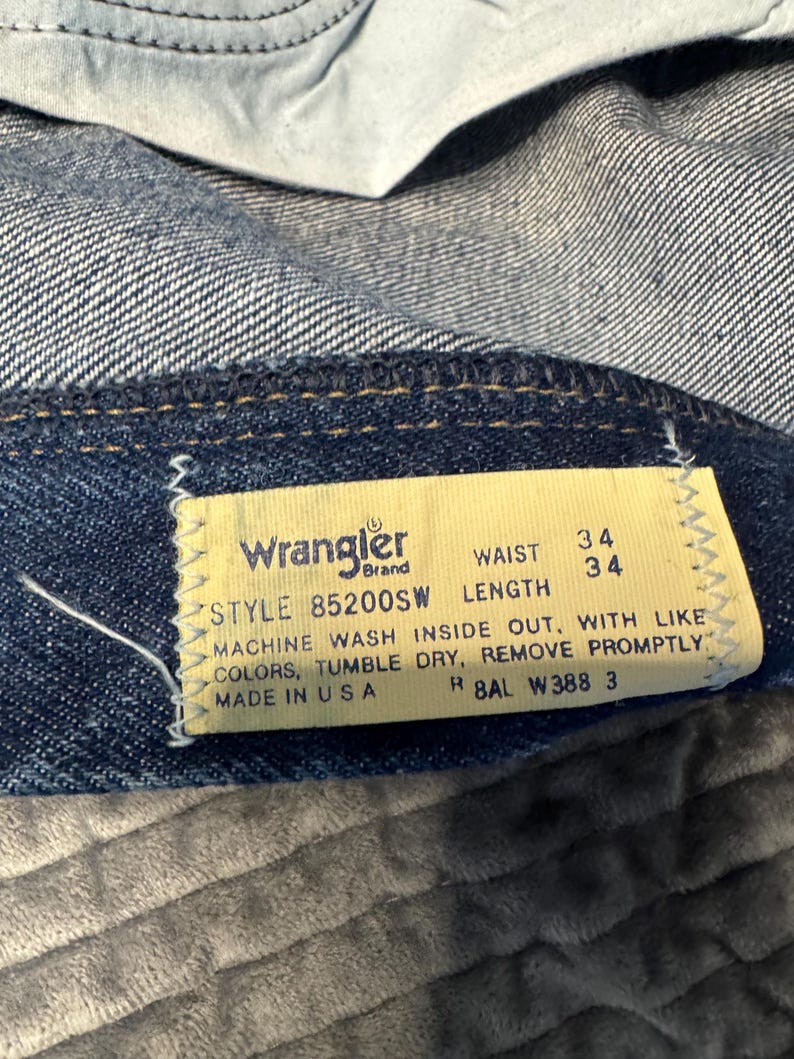 K&ouml;nnte beinhalten: Nahaufnahme eines Jeansetiketts der Marke Wrangler. Das gelbe Etikett mit schwarzem Text zeigt eine Bundweite von 86 cm und eine L&auml;nge von 86 cm an. Das Etikett enth&auml;lt auch Waschanweisungen und die Modellnummer 85200SW. Die Jeans ist blau.