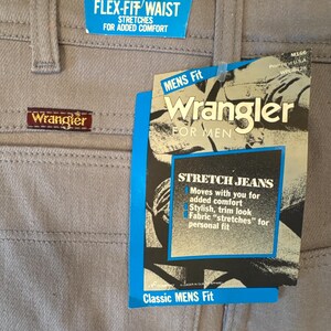 May include: Close-up of light gray Wrangler jeans with tags. The tags read "FLEX-FIT WAIST" and "STRETCH JEANS". The jeans have a classic fit and are designed for added comfort.