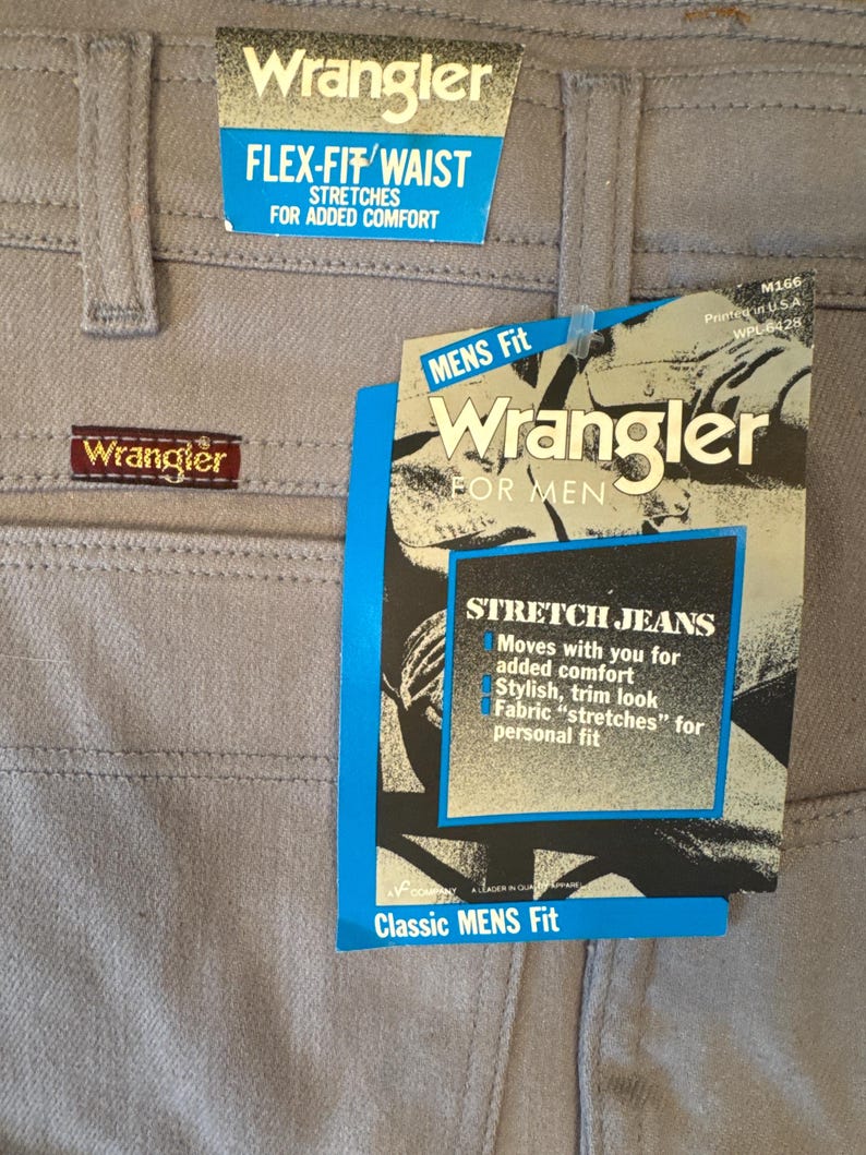 May include: Close-up of light gray Wrangler jeans with tags. The tags read "FLEX-FIT WAIST" and "STRETCH JEANS". The jeans have a classic fit and are designed for added comfort.