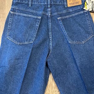 May include: Back view of blue denim Wrangler jeans. The jeans have two back pockets and a brown leather patch with the Wrangler logo. The jeans are folded and displayed on a wooden surface.