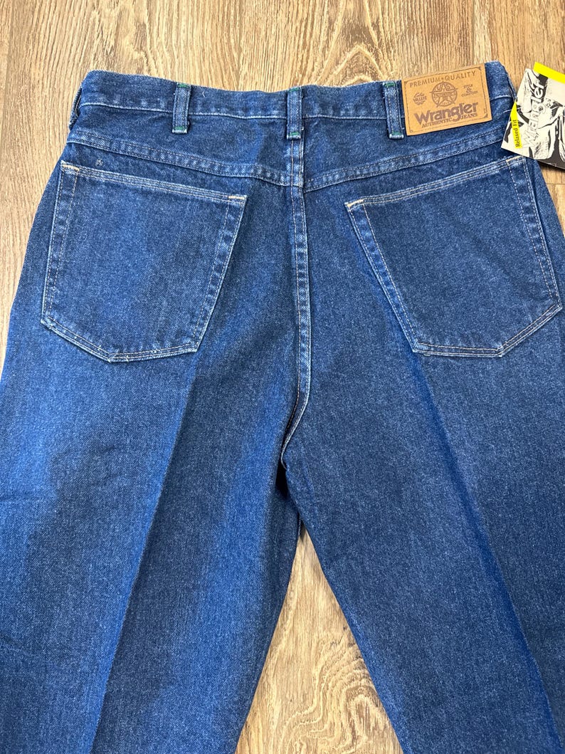 May include: Back view of blue denim Wrangler jeans. The jeans have two back pockets and a brown leather patch with the Wrangler logo. The jeans are folded and displayed on a wooden surface.