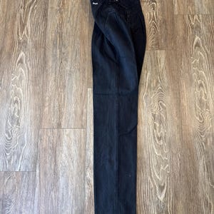 May include: Dark wash denim jeans with a high waist and straight leg. The jeans are folded and lying on a wooden surface. The jeans have a button and zip closure and a small tag on the back pocket.