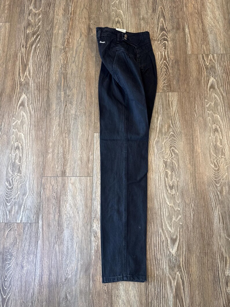 May include: Dark wash denim jeans with a high waist and straight leg. The jeans are folded and lying on a wooden surface. The jeans have a button and zip closure and a small tag on the back pocket.