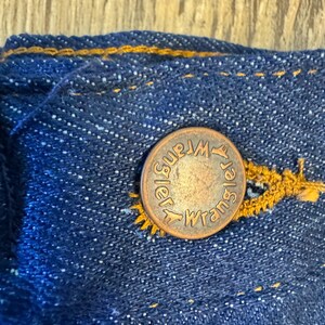 May include: Close-up of a copper-colored button with the word "Wrangler" embossed on it, attached to a blue denim fabric. The button is secured with yellow stitching, and the denim has a textured appearance.