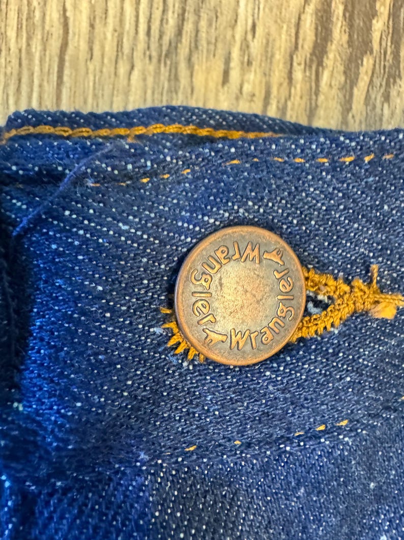 May include: Close-up of a copper-colored button with the word "Wrangler" embossed on it, attached to a blue denim fabric. The button is secured with yellow stitching, and the denim has a textured appearance.