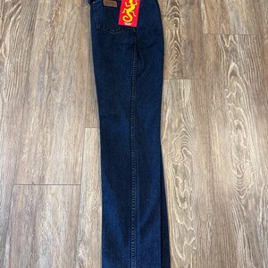 May include: Dark blue denim jeans with a brown leather patch and a red tag with the word "RIATA" and a yellow lizard graphic. The jeans are folded and lying on a wooden surface.